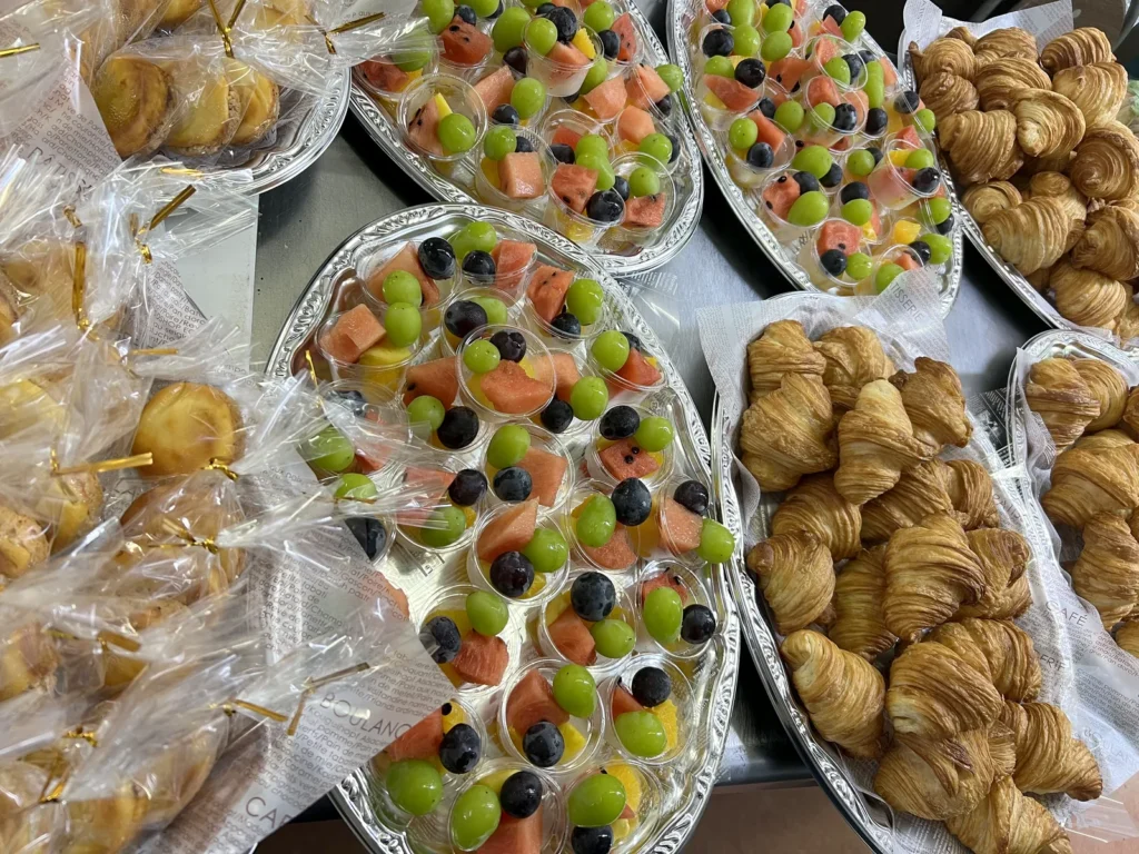種類ごとに大皿に並べたイベント用の軽食（フルーツカップ・クロワッサン・個包装サンドイッチ）